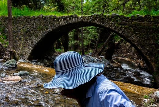 A girl in a wide-brimmed hat drinks water from a stream next to a medieval Venetian bridge in the center of the island of Cyprus.