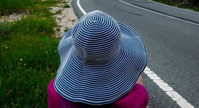A girl in a wide-brimmed striped hat is waiting for transport on the side of the road