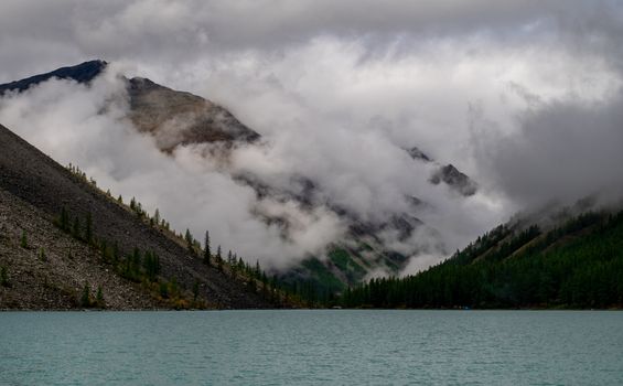Shavlinskoye lake in the Altai Republic on the background of mountain peaks shrouded in fog.