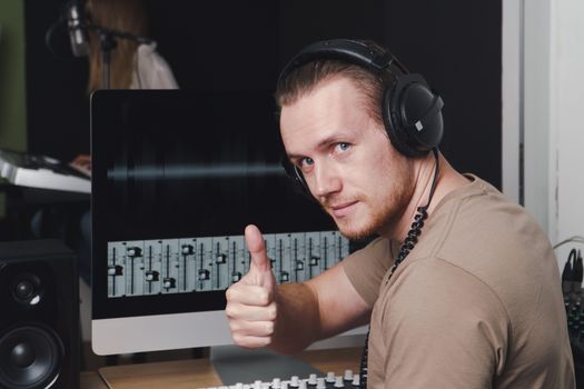portrait young, adult Caucasian man wearing a headphone, raising his hand, and smiling. Sound producer operate on computer monitor and Audio mixer panels in the recording studio.