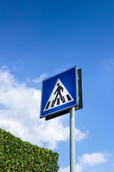 Pedestrian crossing sign with blue sky in background and green hedge.