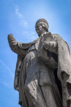 Statue of a pope with mutilated hands, against a blue sky and sunlight.