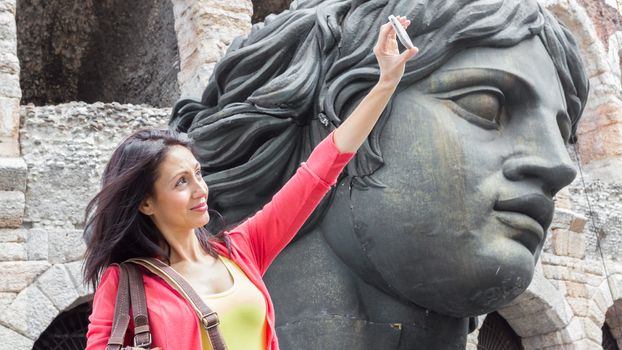 Young woman traveler making selfie photo with details of the Arena in Verona (Italy) and scenic decorations on background. Shallow DOF.