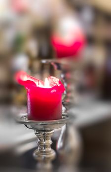 Close-up detail of red candle in silver candlestick lighting up a church. On Defocused blurry background they can be seen other red candle. Symbol of religion, prayer and peace.