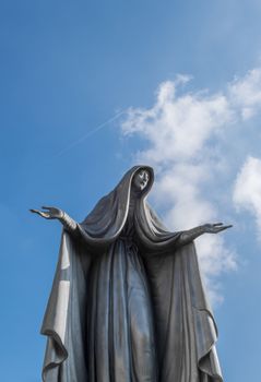 Statue of Virgin Mary in bronze, in a blue sky background with white clouds, with outstretched arms.