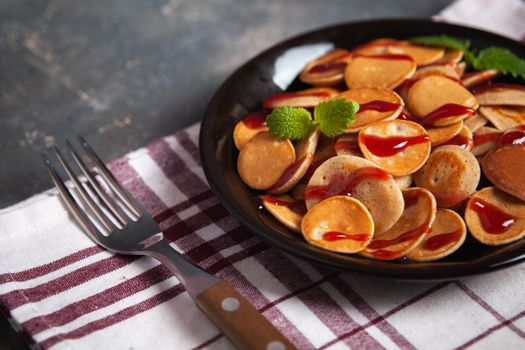 Trendy homemade breakfast, pancake cereal, mini pancakes on dark plate on grunge gray concrete background with check towel and fork, leaves of lemon balm or peppermint and cherry jam. Space for text.
