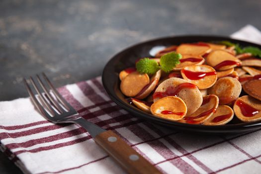 Trendy homemade breakfast, pancake cereal, mini pancakes on dark plate on grunge gray concrete background with check towel and fork, leaves of lemon balm or peppermint and cherry jam. Space for text.