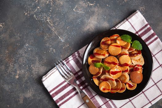Trendy homemade breakfast, pancake cereal, mini pancakes on dark plate on grunge gray concrete background with check towel and fork, leaves of lemon balm or peppermint and cherry jam. Space for text.