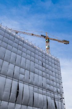 Building facade renovation with cellophane as protection and crane, against a blue sky with white clouds.