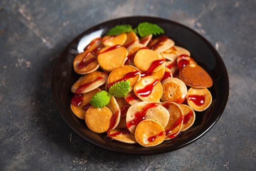 Trendy homemade breakfast, pancake cereal, mini pancakes on dark plate on grunge gray concrete background, leaves of lemon balm or peppermint and cherry jam. Space for text.
