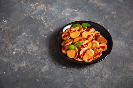 Trendy homemade breakfast, pancake cereal, mini pancakes on dark plate on grunge gray concrete background, leaves of lemon balm or peppermint and cherry jam. Space for text.
