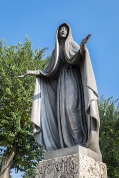 Statue of Virgin Mary in bronze, in a blue sky background with white clouds and tree, with outstretched arms.