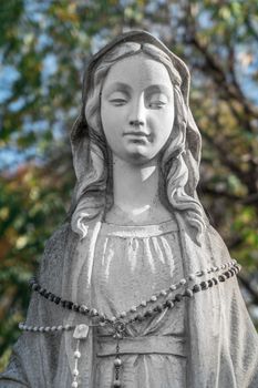 Virgin Mary Statue. On defocused blurry background some green trees. Ideal for concepts and festivity.