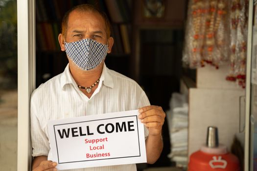 Small business owner in medical mask holding Wellcome notice infront of door after store reopening during coronavirus or covid-19 - concept of support local business and restart work after pandemic