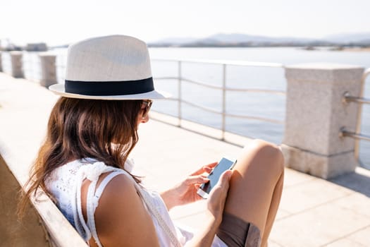 Caucasian young woman with white hat sitting on a bench of a sidewalk by the sea using her smartphone - Single woman outdoor connected with new social technology