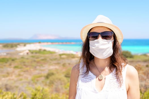 Caucasian young woman posing for a picture with Coronavirus protection mask in summer vacation with bright and colored tropical sea in the background