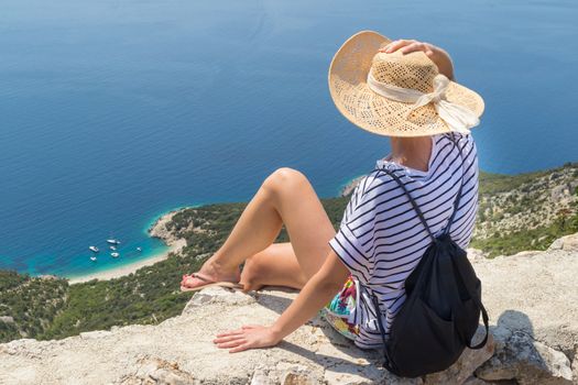 Active sporty woman on summer vacations sitting on old stone wall at Lubenice village, wearing straw hat and beach backpack enjoying beautiful coastal view of Cres island, Croatia.