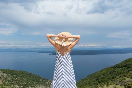 Rear view of young woman wearing striped summer dress and straw hat standing in super bloom of wildflowers, relaxing while enjoing beautiful view of Adriatic sea nature, Croatia.