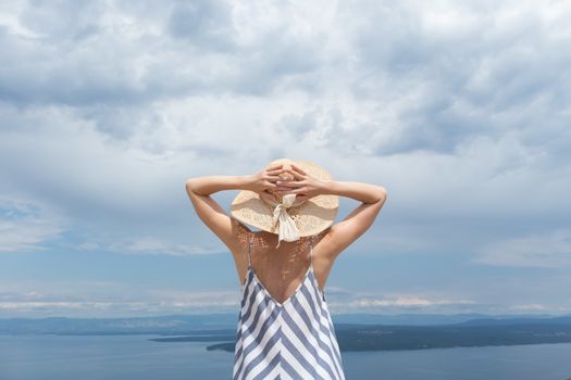 Rear view of young woman wearing striped summer dress and straw hat standing in super bloom of wildflowers, relaxing while enjoing beautiful view of Adriatic sea nature, Croatia.