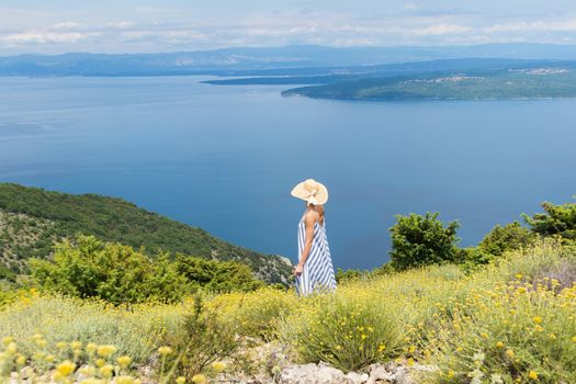 Rear view of young woman wearing striped summer dress and straw hat standing in super bloom of wildflowers, relaxing while enjoing beautiful view of Adriatic sea nature, Croatia.