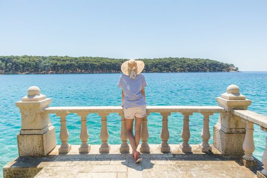 Rear view of woman wearing straw summer hat ,leaning against elegant old stone fence of coastal villa, relaxing while looking at blue Adriatic sea, on Losinj island Croatia.