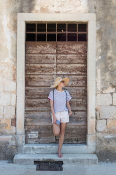 Beautiful young female tourist woman standing in front of vinatage wooden door and textured stone wall at old Mediterranean town, smiling, holding, smart phone to network on vacationes.