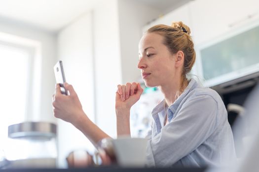 Young woman indoors at home kitchen using social media apps on phone for video chatting and stying connected with her loved ones. Stay at home, social distancing lifestyle.