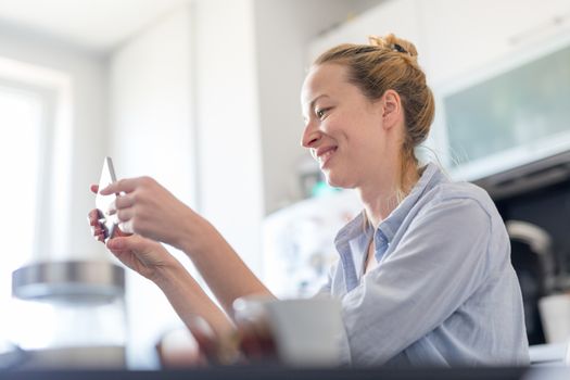 Young smiling cheerful woman indoors at home kitchen using social media on phone for video chatting and stying connected with her loved ones. Stay at home, social distancing lifestyle.