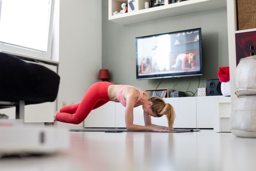 Attractive sporty woman working out at home, doing pilates exercise in front of television in her living room. Social distancing. Stay healthy and stay at home during corona virus pandemic.