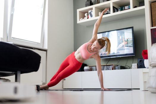 Attractive sporty woman working out at home, doing pilates exercise in front of television in her living room. Social distancing. Stay healthy and stay at home during corona virus pandemic.