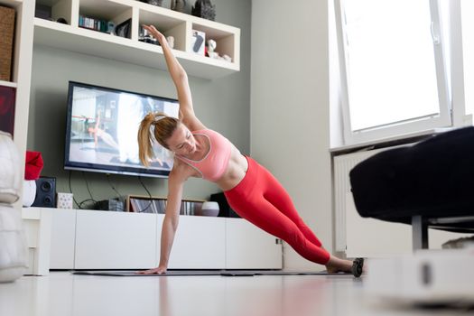 Attractive sporty woman working out at home, doing pilates exercise in front of television in her living room. Social distancing. Stay healthy and stay at home during corona virus pandemic.