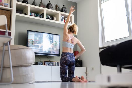 Attractive sporty woman working out at home, doing pilates exercise in front of television in her living room. Social distancing. Stay healthy and stay at home during corona virus pandemic.