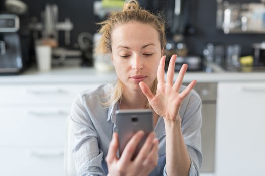Young woman indoors at home kitchen using social media apps on phone for video chatting and stying connected with her loved ones. Stay at home, social distancing lifestyle.