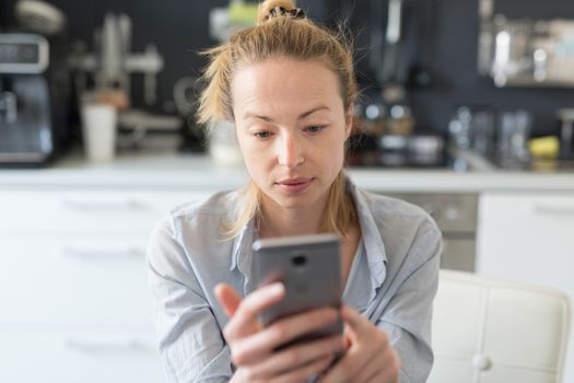 Worried woman indoors at home kitchen using social media apps on phone for video chatting and stying connected with her loved ones. Stay at home, social distancing lifestyle.