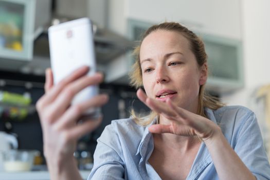 Young woman indoors at home kitchen using social media apps on phone for video chatting and stying connected with her loved ones. Stay at home, social distancing lifestyle.