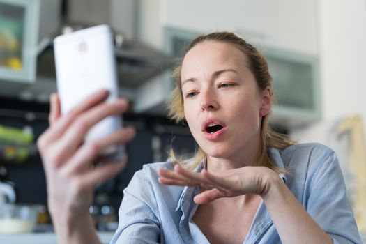 Young woman indoors at home kitchen using social media apps on phone for video chatting and stying connected with her loved ones. Stay at home, social distancing lifestyle.