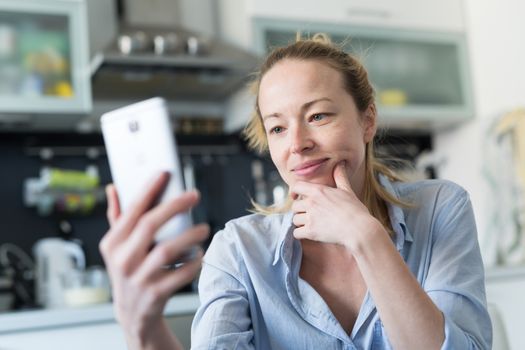 Young smiling cheerful woman indoors at home kitchen using social media apps on phone for video chatting and stying connected with her loved ones. Stay at home, social distancing lifestyle.