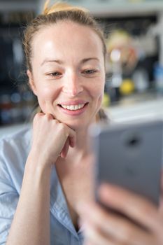 Young smiling cheerful woman indoors at home kitchen using social media apps on phone for video chatting and stying connected with her loved ones. Stay at home, social distancing lifestyle.