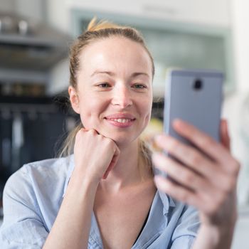 Young smiling cheerful woman indoors at home kitchen using social media apps on phone for video chatting and stying connected with her loved ones. Stay at home, social distancing lifestyle.