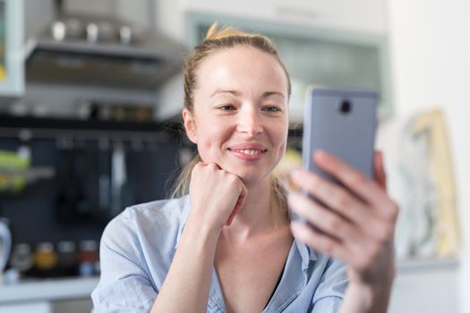 Young smiling cheerful woman indoors at home kitchen using social media apps on phone for video chatting and stying connected with her loved ones. Stay at home, social distancing lifestyle.