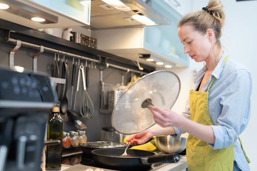 Stay at home housewife woman cooking in kitchen, stir frying dish in a saucepan, preparing food for family dinner. Blonde caucasian model in her 30is, real cooking not looking in camera.