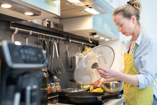 Stay at home housewife woman cooking in kitchen, stir frying dish in a saucepan, preparing food for family dinner. Blonde caucasian model in her 30is, real cooking not looking in camera.