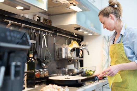 Stay at home housewife woman cooking in kitchen, stir frying dish in a saucepan, preparing food for family dinner. Blonde caucasian model in her 30is, real cooking not looking in camera.
