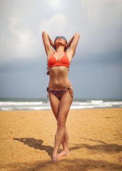 Young woman in red swimsuit and sunglasses, standing on the beach holding her hands behind head, looking up into the sun. Kalutara beach, Sri Lanka