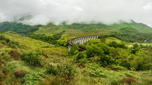 Glenfinnan railway viaduct (location from Harry Potter movie), on overcast day with grey sky and lot of green grass and trees around. Inverness, Scotland