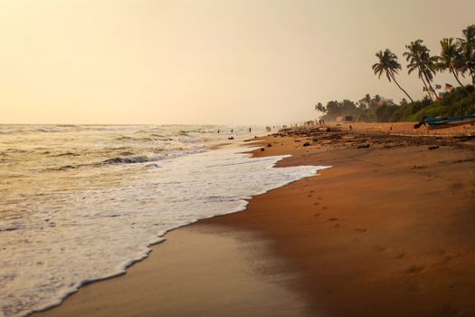 Empty beach during afternoon sun light, sand wet from waves, with silhouettes of people playing in background. Kalutara, Sri Lanka