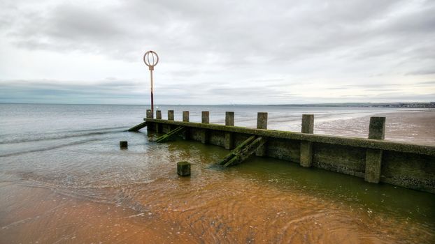 Old wooden groyne structure covered with green algae on Portobello beach during low tide with North sea in the background shot on overcast day. Edinburgh, Scotland.