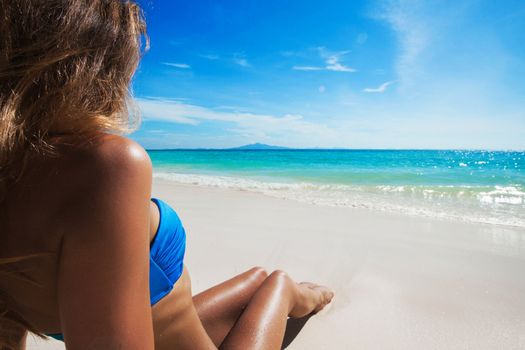 Beautiful tan woman in bikini lies on white sand beach looking on the sea, back view