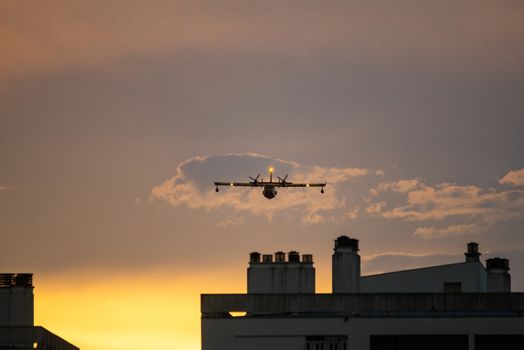 Amphibious water bomber in flight at sunset, airplane flying low over buildings, in France.