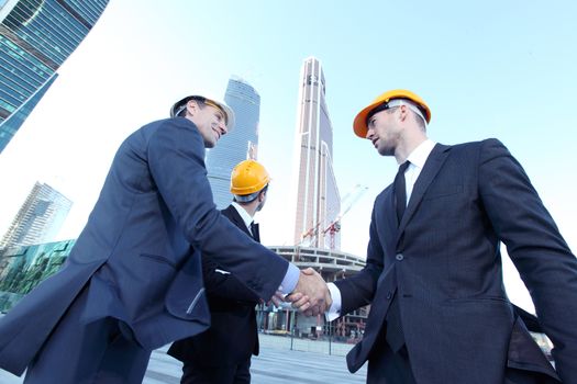 Architects wearing helmets shaking hands at skyscrapers background